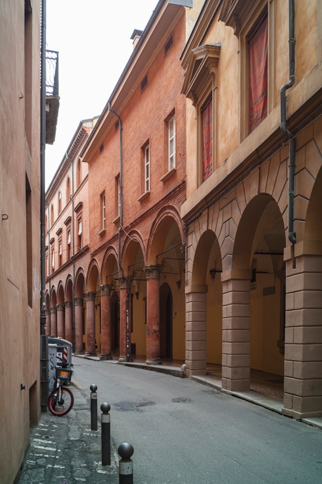 Una strada stretta e affascinante a Bologna, fiancheggiata da edifici storici con i caratteristici portici. Le arcate in mattoni rossi e pietra creano un percorso coperto, tipico dell'architettura bolognese, che conferisce alla via un'atmosfera unica e accogliente.